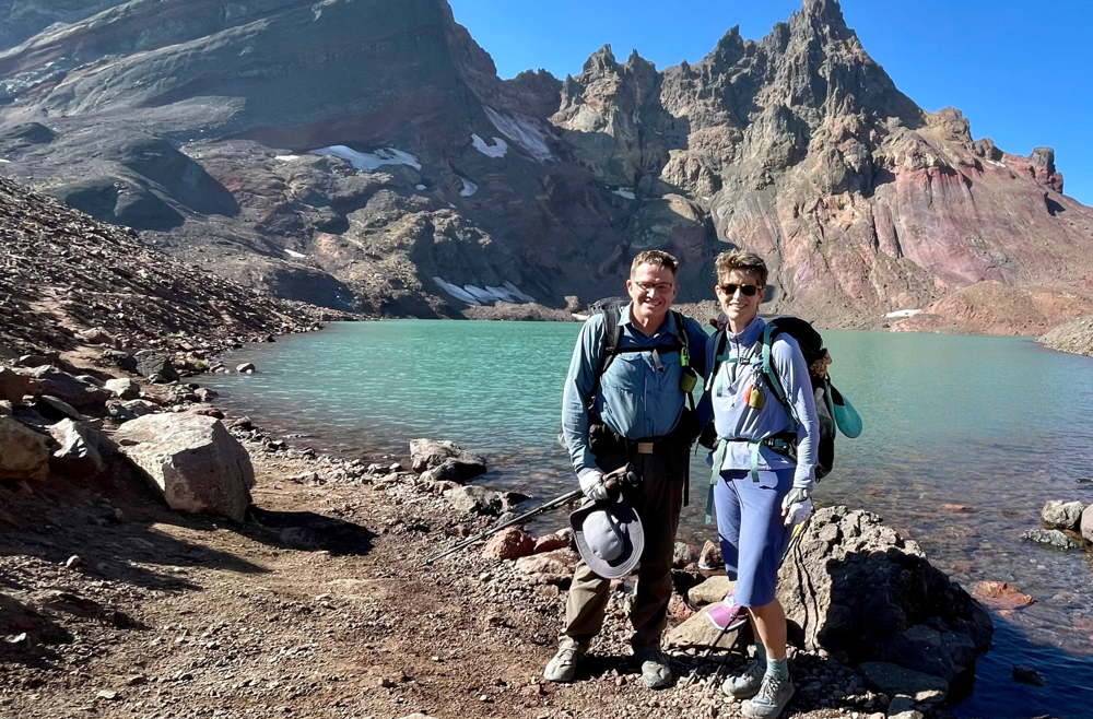 A couple in hiking gear pose in front of a mountain lake. 